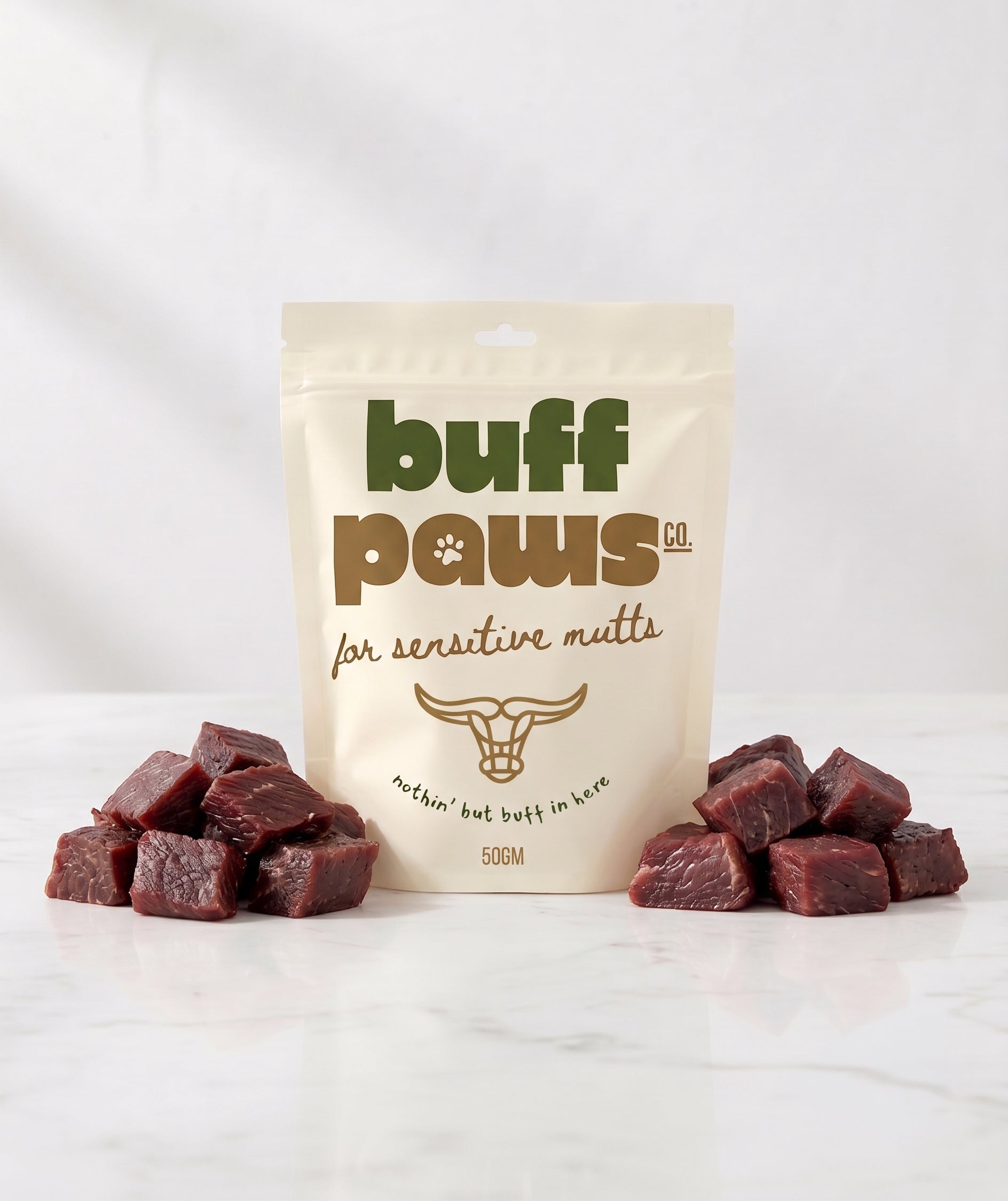 A 50g stand-up pouch of Buff Paws Co. buffalo jerky dog treats on a white marble surface, flanked by two small piles of dark red cured meat cubes.