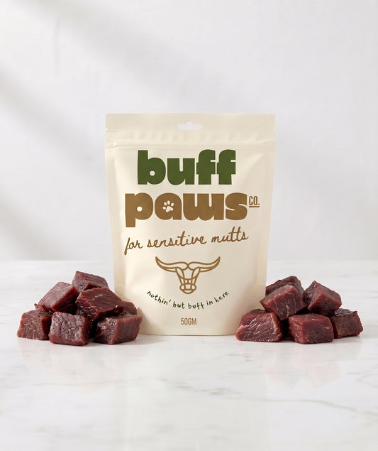 A 50g stand-up pouch of Buff Paws Co. buffalo jerky dog treats on a white marble surface, flanked by two small piles of dark red cured meat cubes.
