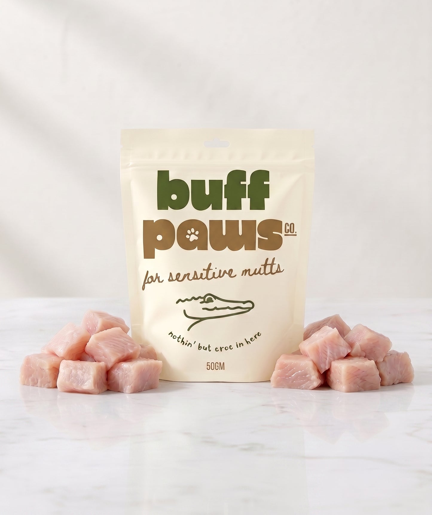 A stand-up pouch of buff paws natural raw meat treats for sensitive mutts, displayed on a white marble surface with pink raw crocodile meat cubes arranged on both sides.