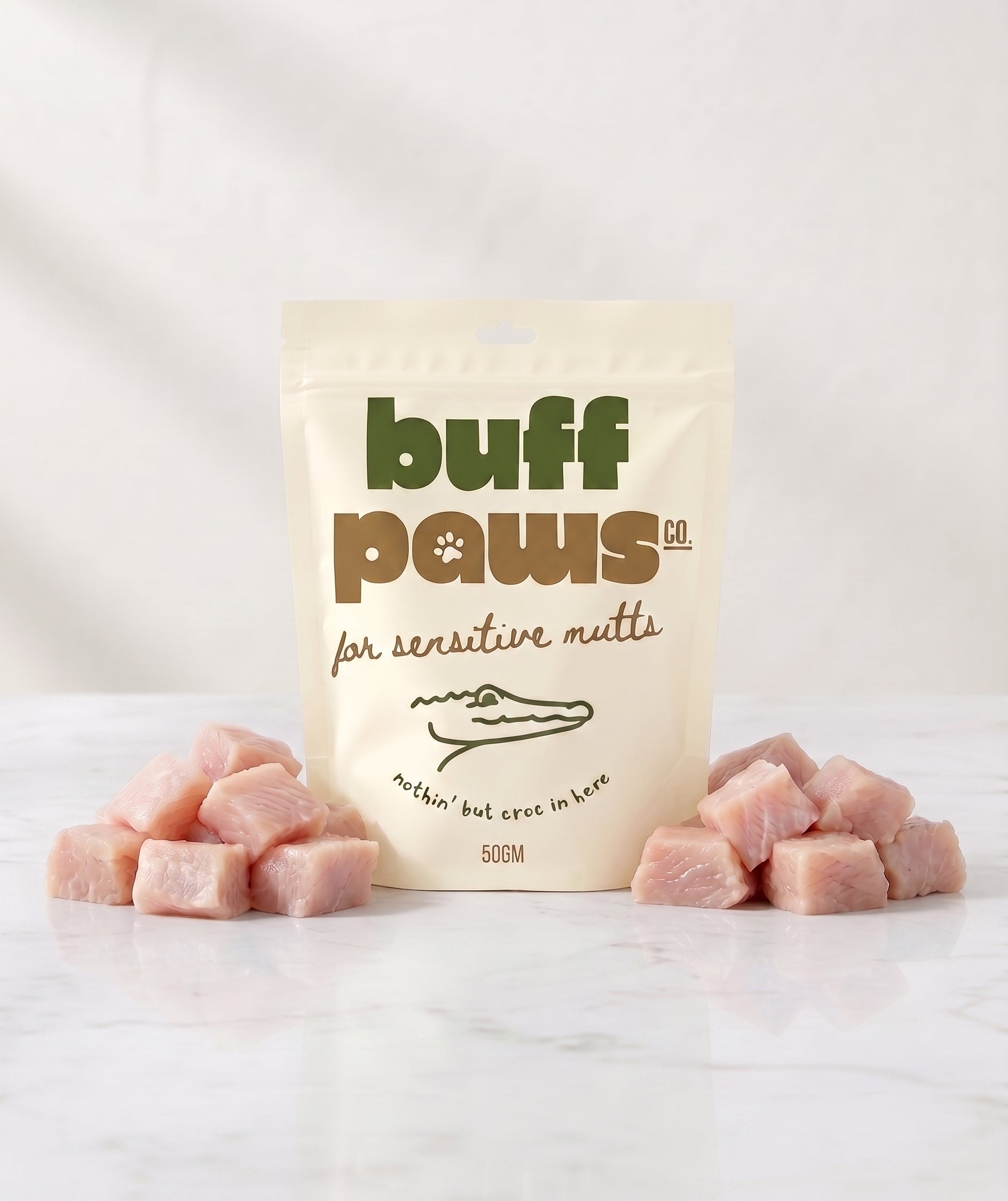 A stand-up pouch of buff paws natural raw meat treats for sensitive mutts, displayed on a white marble surface with pink raw crocodile meat cubes arranged on both sides.