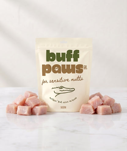 A stand-up pouch of buff paws natural raw meat treats for sensitive mutts, displayed on a white marble surface with pink raw crocodile meat cubes arranged on both sides.