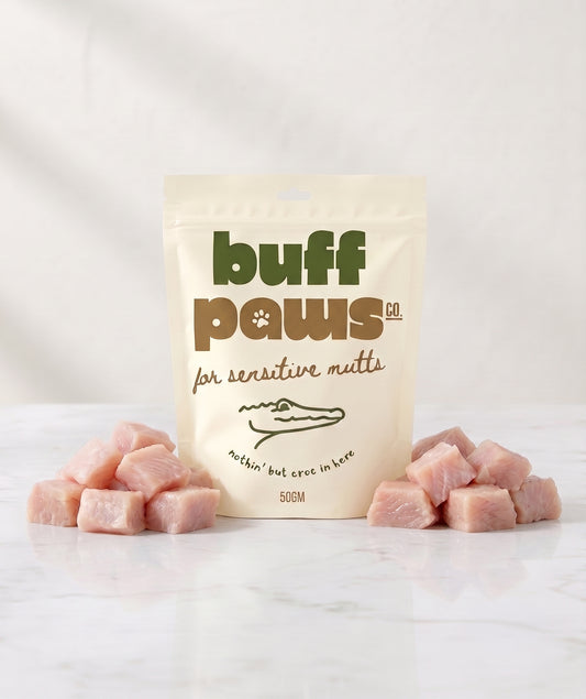 A stand-up pouch of buff paws natural raw meat treats for sensitive mutts, displayed on a white marble surface with pink raw crocodile meat cubes arranged on both sides.