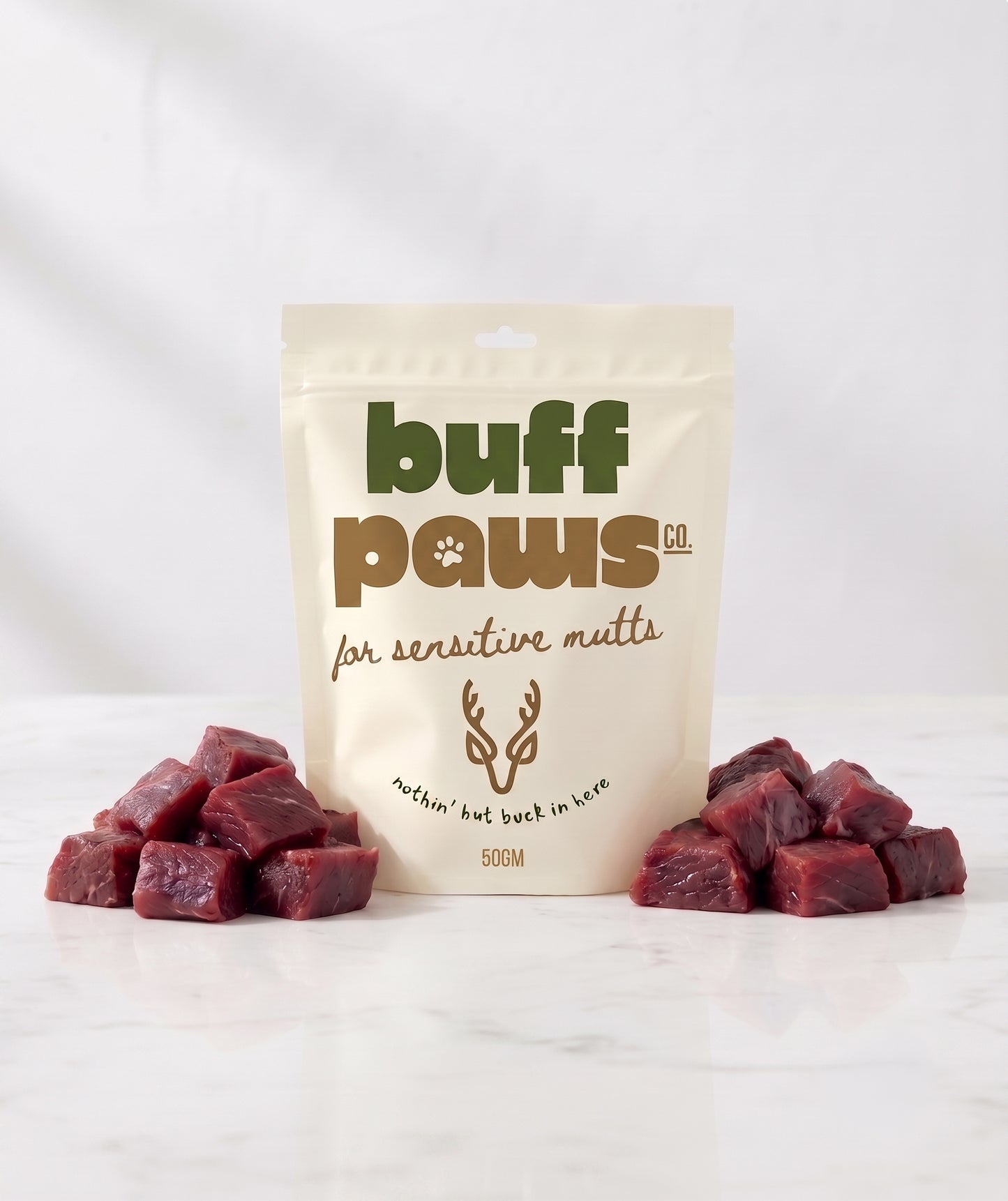 A stand-up pouch of buff paws dog treats for sensitive guts, flanked by two small piles of raw Venison cube treats on a white marble surface against a white background.
