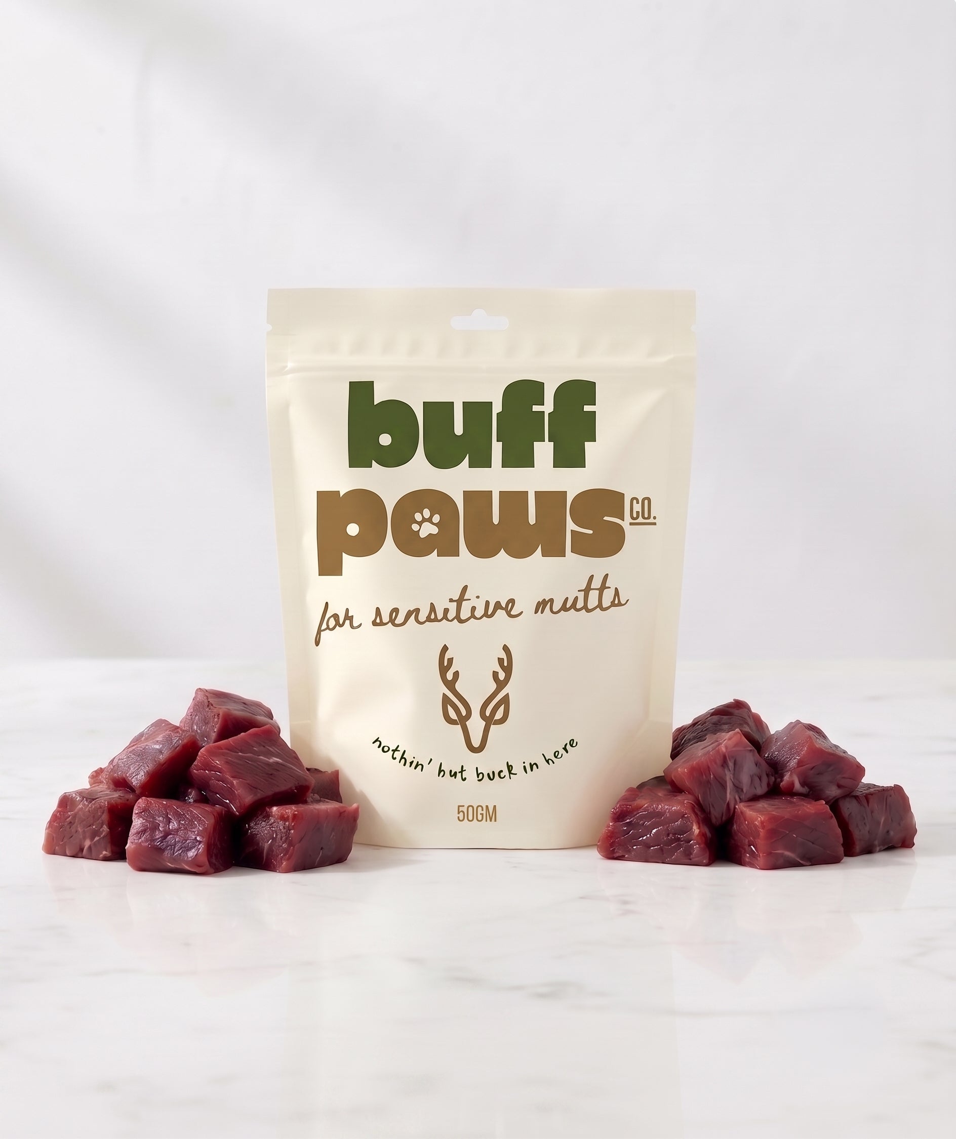 A stand-up pouch of buff paws dog treats for sensitive guts, flanked by two small piles of raw Venison cube treats on a white marble surface against a white background.