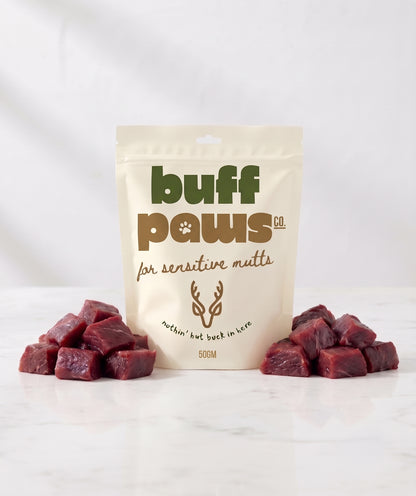 A stand-up pouch of buff paws dog treats for sensitive guts, flanked by two small piles of raw Venison cube treats on a white marble surface against a white background.