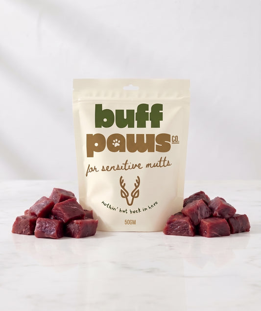 A stand-up pouch of buff paws dog treats for sensitive guts, flanked by two small piles of raw Venison cube treats on a white marble surface against a white background.